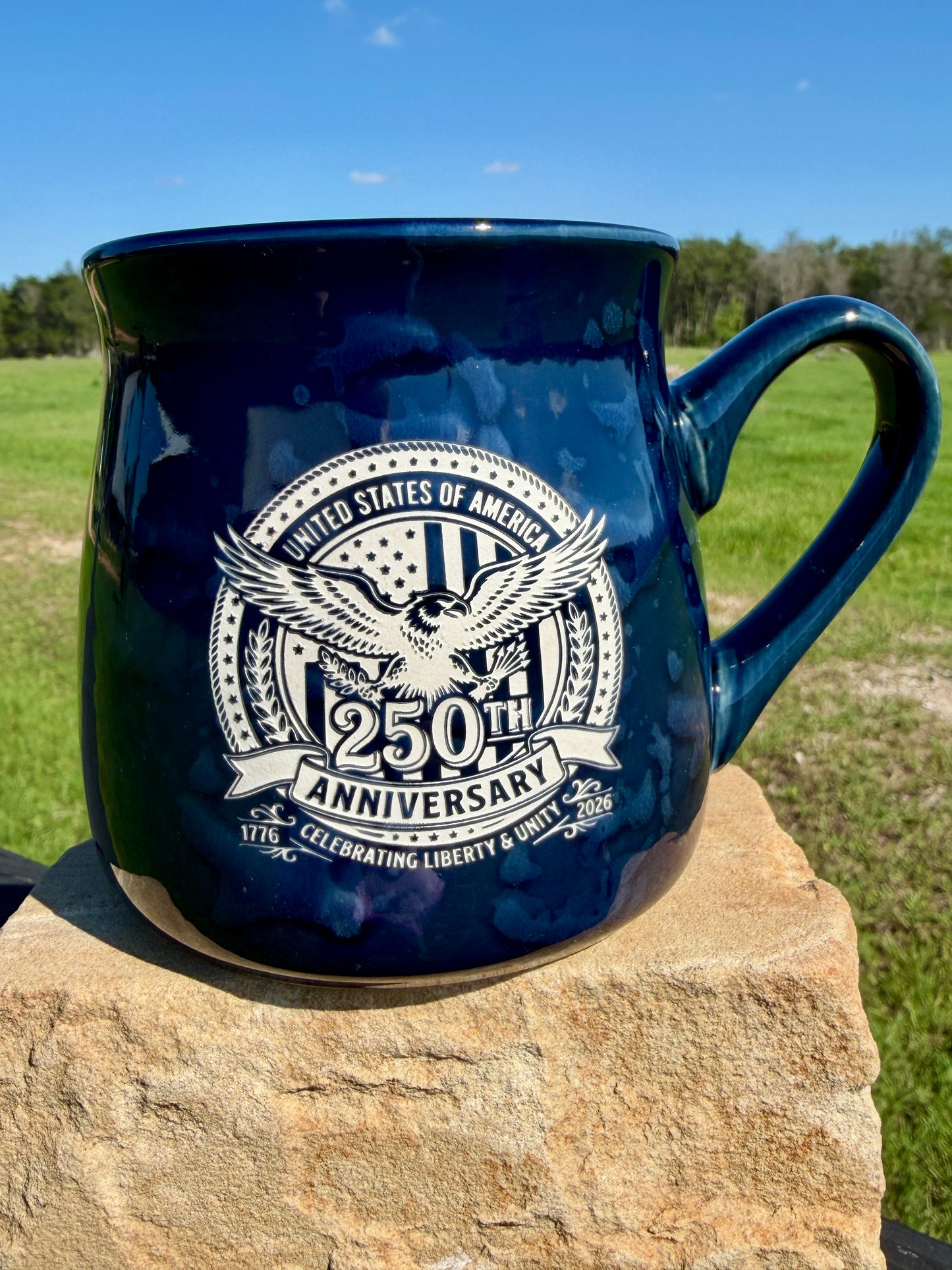 Navy blue mug with 250th Anniversary design on a stone surface with grass and trees in the background