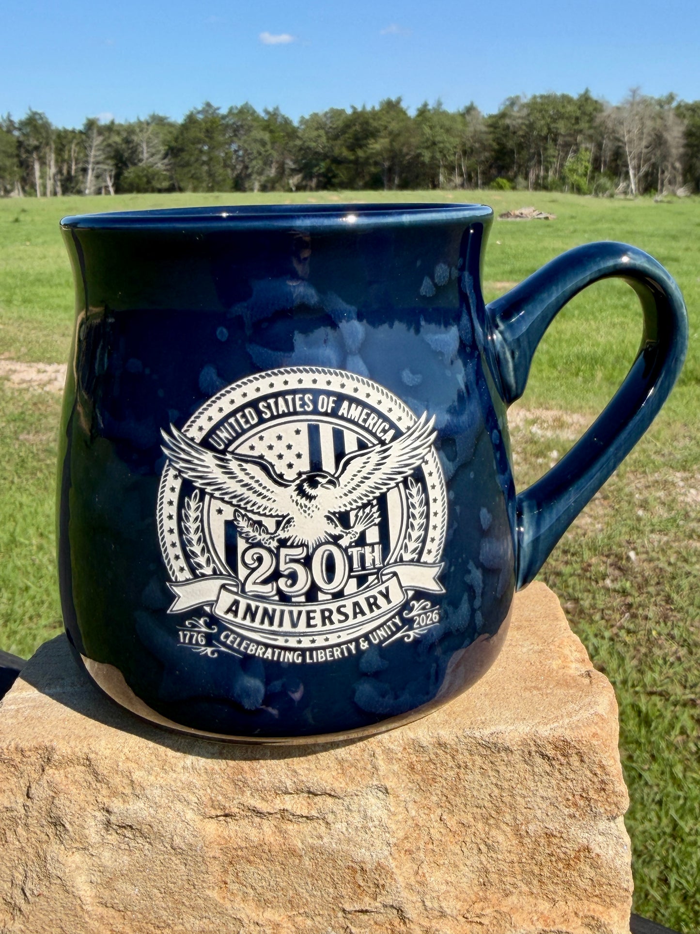 Blue mug with 250th Anniversary design on a stone surface with a grassy field and trees in the background