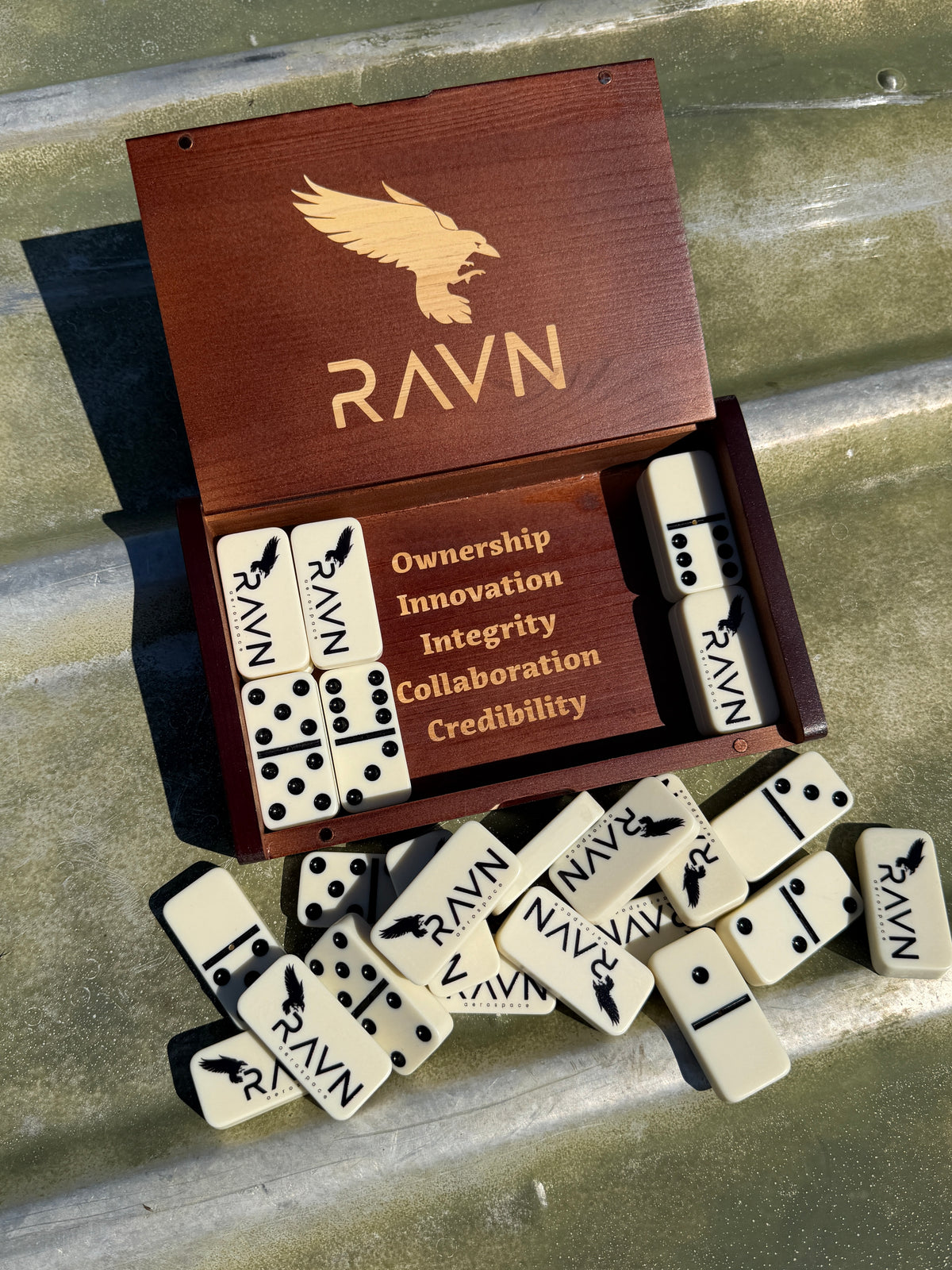 A wooden game box labeled RAVN with cream-colored domino tiles printed with black text and symbols, arranged on a corrugated metal surface.