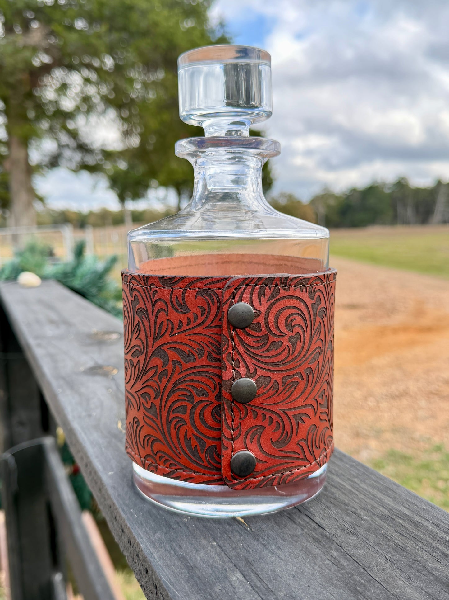 Glass decanter with a brown leather wrap featuring metal studs on a wooden surface with a natural background.